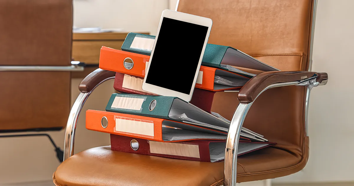 Das Bild zeigt einen Stuhl, auf dem ein Haufen Aktenordner liegt. Oben auf dem Haufen liegt ein Tablet.