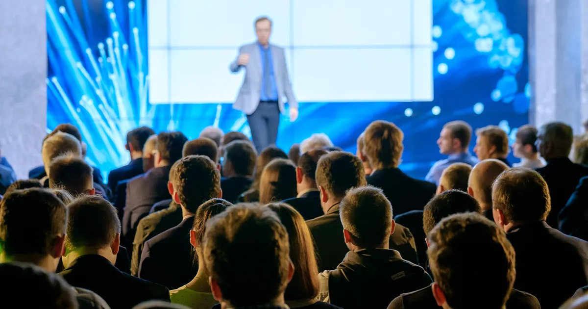 Ein Publikum in einer Messehalle lauscht den Ausführungen eines Speakers.